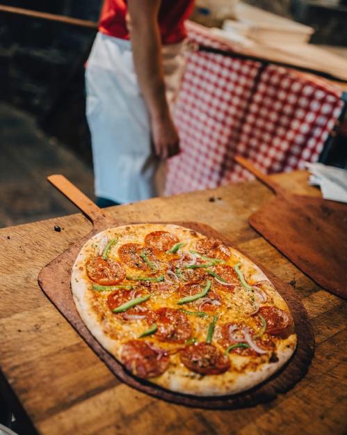 A freshly baked pizza with pepperoni, green peppers, and onions on a wooden peel, with a chef in the background.