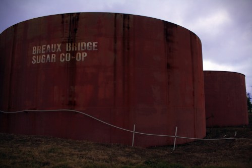 Two large, rusted storage tanks with "Breaux Bridge Sugar Co-Op" painted on the side, set against a cloudy sky.