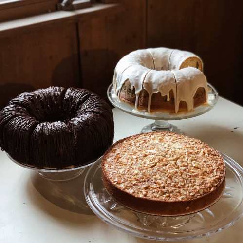 Three cakes on glass stands: a chocolate bundt, a glazed bundt, and a coconut cake, all set on a rustic table.