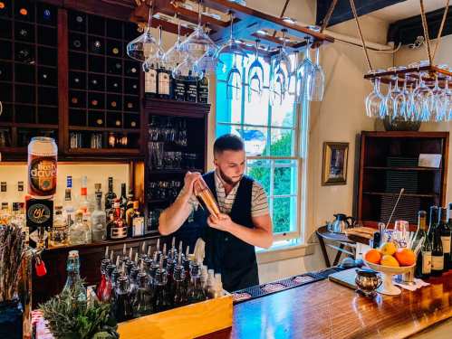 A bartender shakes a cocktail at a well-stocked bar with bottles and glasses in a cozy setting.