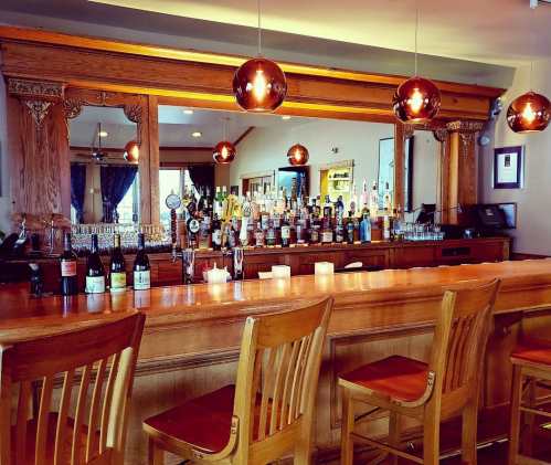 A wooden bar with empty stools, illuminated by pendant lights, showcasing a variety of liquor bottles in the background.