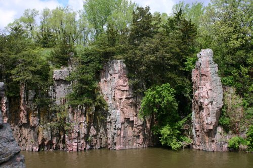Rocky cliffs rise above a calm river, surrounded by lush green trees under a partly cloudy sky.