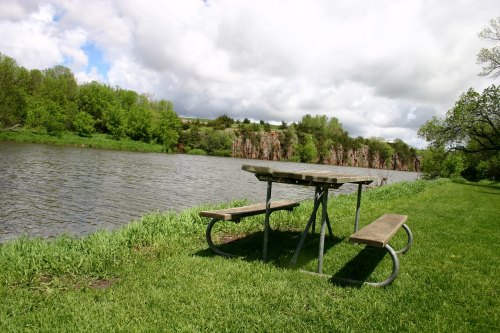 A picnic table by a calm river, surrounded by lush greenery and rocky cliffs under a cloudy sky.