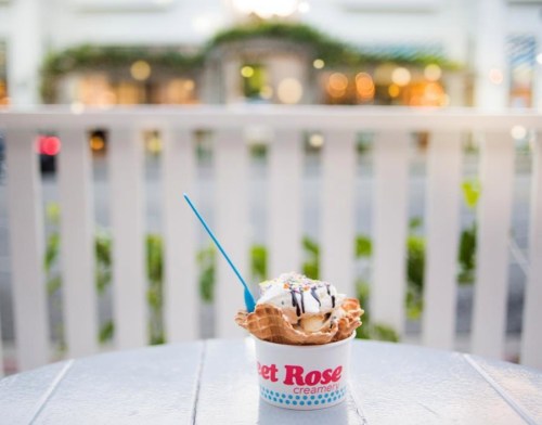 A cup of ice cream with a waffle cone, topped with chocolate drizzle and sprinkles, sits on a table.