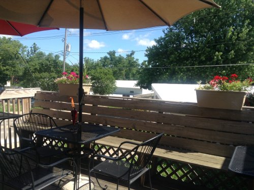 A cozy outdoor seating area with a table, chairs, and potted flowers under a large umbrella, surrounded by greenery.