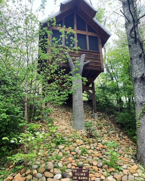 A treehouse perched on a rocky slope, surrounded by greenery, with a sign reading "Do Not Climb on Banks."