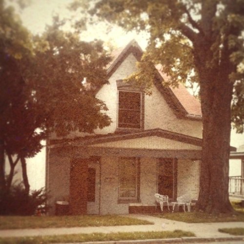 A vintage-style house surrounded by trees, featuring a porch and a small garden in front.