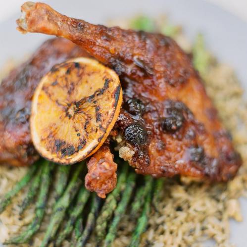 A plate featuring crispy chicken leg, garnished with a dried orange slice, served on a bed of rice and asparagus.