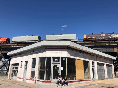 A small, boarded-up building with two people sitting outside, under a blue sky and a train passing overhead.
