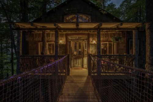 A cozy treehouse with warm lighting, featuring a wooden bridge and surrounded by lush trees at dusk.