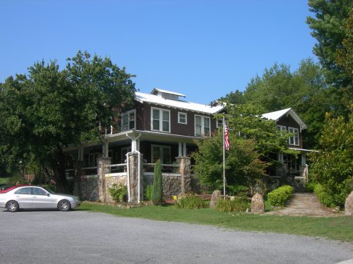 A large, two-story house with a stone facade, surrounded by trees and a grassy area, under a clear blue sky.