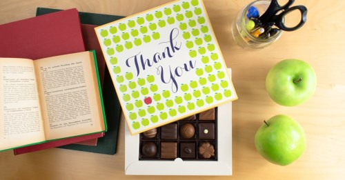 A "Thank You" chocolate box on a table with green apples, an open book, and a jar of pens and scissors.
