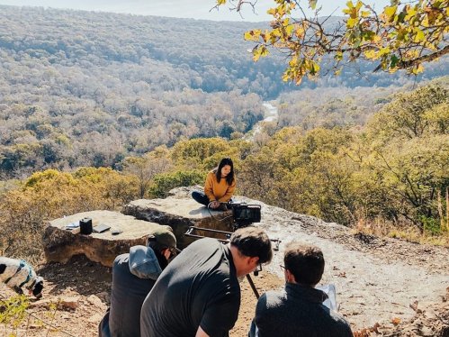 A woman sits on a rock ledge while a group of people films her against a scenic, wooded landscape.