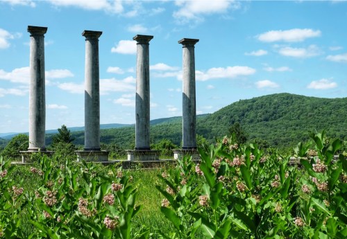 Four tall stone columns stand amidst lush greenery and rolling hills under a blue sky with fluffy clouds.
