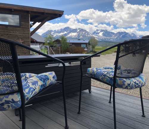 Two chairs with floral cushions face a scenic view of mountains under a partly cloudy sky, from a wooden deck.