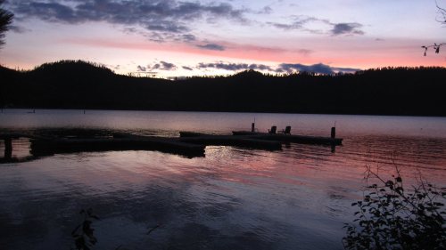 A serene lake at sunset, with silhouettes of a dock and mountains reflecting in the water.