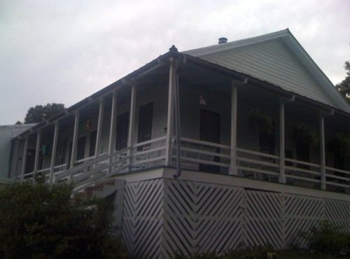 A light blue house with a porch, featuring white railings and a sloped roof, set against a cloudy sky.