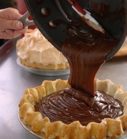 A hand pours rich chocolate filling into a pie crust, with a meringue pie in the background.