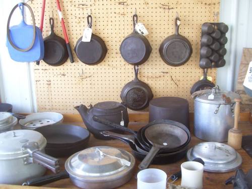 A collection of vintage cookware, including cast iron pans, a kettle, and various pots, displayed on a pegboard.