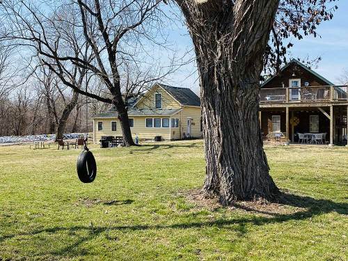 A tire swing hangs from a tree in a grassy yard, with a yellow house and a wooden decked house in the background.