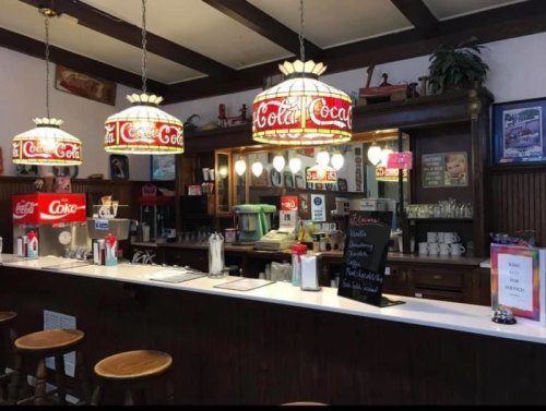 A vintage diner interior featuring a counter with Coca-Cola lamps, stools, and a menu board.
