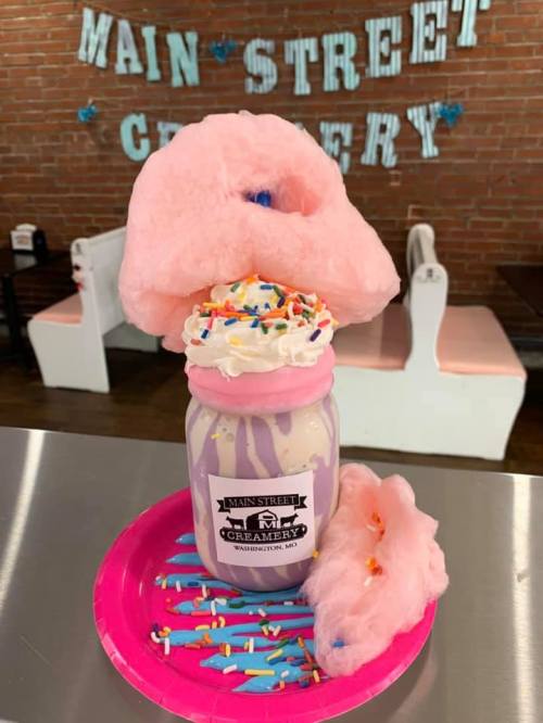 A colorful dessert featuring a milkshake topped with whipped cream, sprinkles, and cotton candy in a jar.