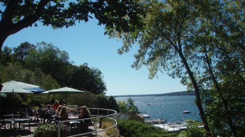 A scenic view of a lake with boats, surrounded by trees and people dining under umbrellas on a sunny day.
