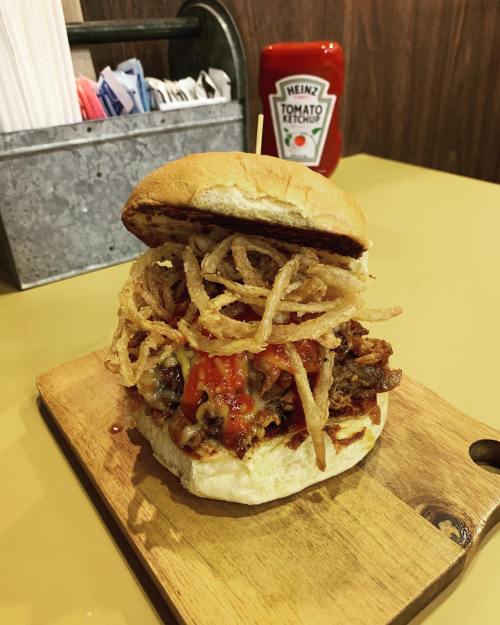 A towering burger topped with crispy onions and sauce, served on a wooden board with a ketchup bottle in the background.