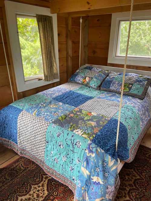 A cozy bedroom with a colorful quilt on a hanging bed, surrounded by wooden walls and windows letting in natural light.