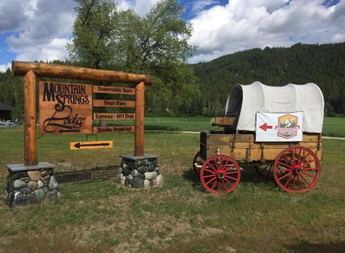 Sign for Mountain Springs Lodge with a wooden wagon nearby, set against a backdrop of green fields and blue skies.
