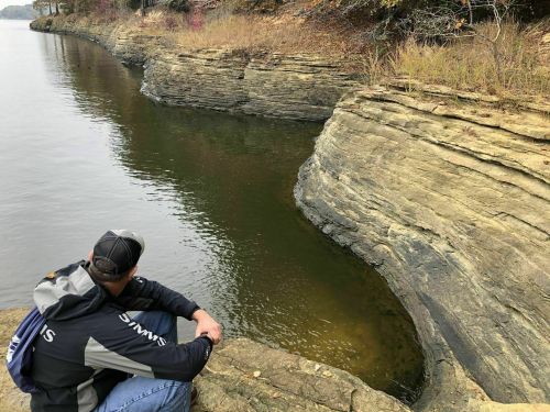 A person sits by a calm lake, observing layered rock formations along the water's edge in a serene natural setting.