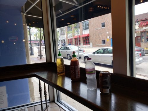 A window view from inside a café, featuring drinks on a ledge and cars parked outside.