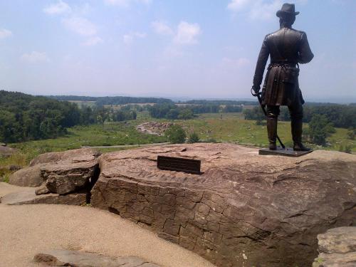 A statue of a soldier stands on a rocky outcrop, overlooking a green landscape under a blue sky.