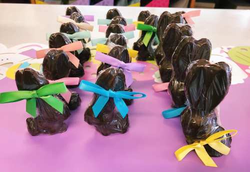 A row of chocolate bunnies wrapped in clear plastic, each adorned with colorful ribbons, on a purple surface.