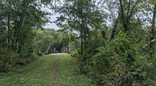 A serene path through greenery leads to a wooden bridge in a peaceful natural setting.