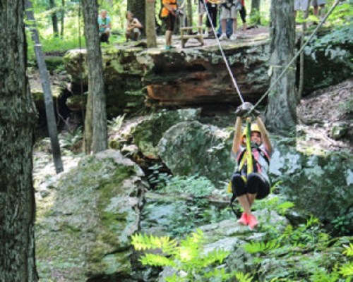 A person zip-lining through a forested area, with spectators watching from a rocky outcrop in the background.