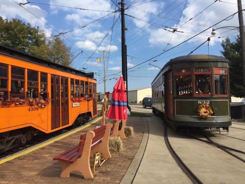 Two vintage streetcars, one orange and one green, at a station with benches and a decorative umbrella under a blue sky.