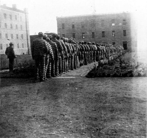 Line of prisoners in striped uniforms walking along a path outside a building, with a guard watching from a distance.