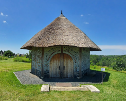 A round building with a thatched roof, set in a grassy area under a clear blue sky.