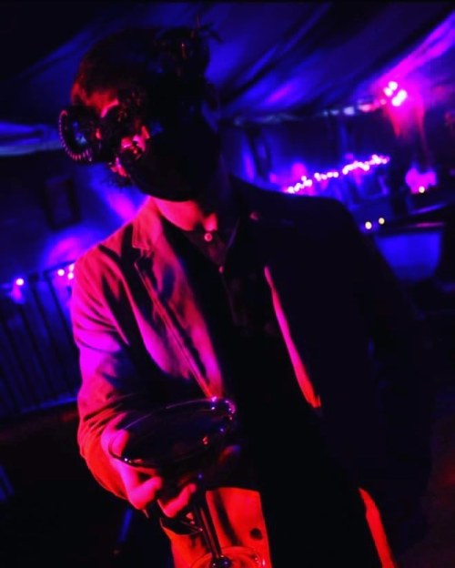 A figure in a dark setting holds a drink, wearing a mask with horns, surrounded by purple and blue lights.