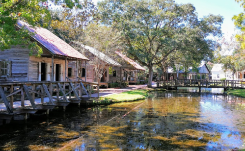 A serene scene of wooden houses by a calm waterway, surrounded by lush trees and greenery.