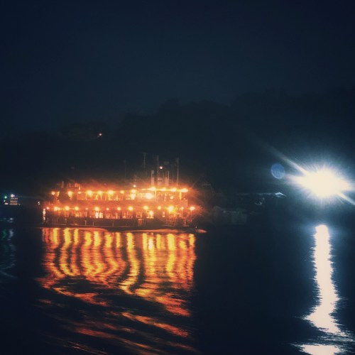 A brightly lit boat reflects on dark water at night, surrounded by a serene landscape.