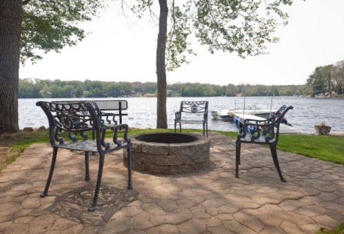 A cozy fire pit area with four chairs by a lake, surrounded by trees and a boat in the background.