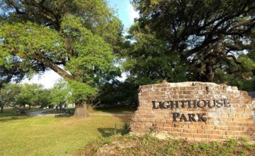A brick sign reading "Lighthouse Park" surrounded by lush green trees and grass on a sunny day.