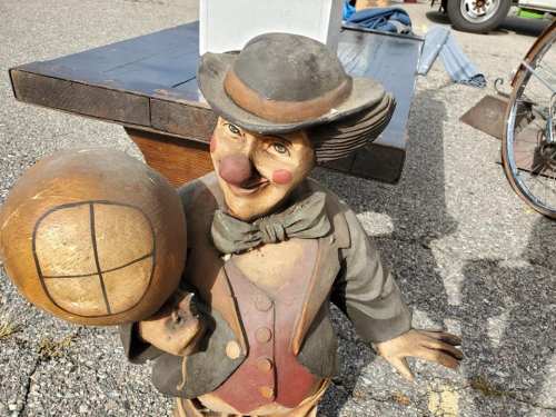 A whimsical wooden clown statue holding a globe, wearing a bow tie and top hat, displayed outdoors on a gravel surface.