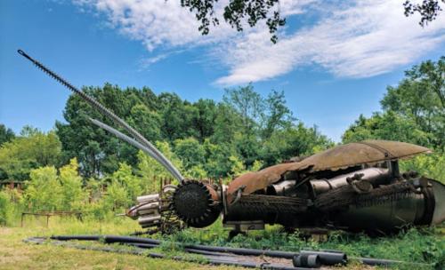 A large, rusted metal sculpture resembling an insect, surrounded by greenery under a blue sky.