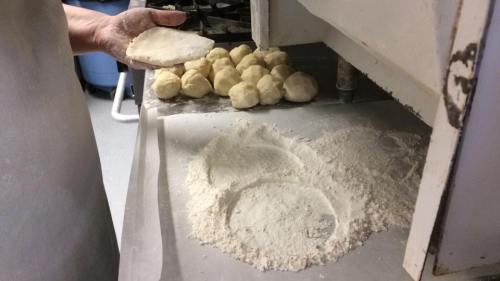 A hand holds a dough piece above a countertop with flour and small dough balls in the background.