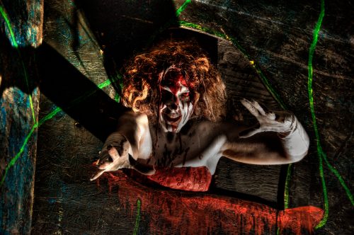 A person with wild hair and dramatic makeup, wearing a red dress, reaches out from a dark, eerie background.