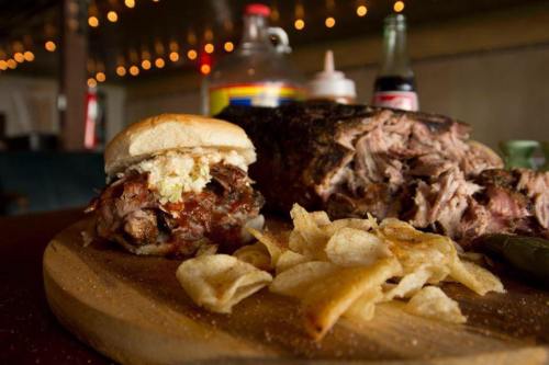 A pulled pork sandwich with coleslaw, served with potato chips on a wooden platter, with barbecue sauce in the background.