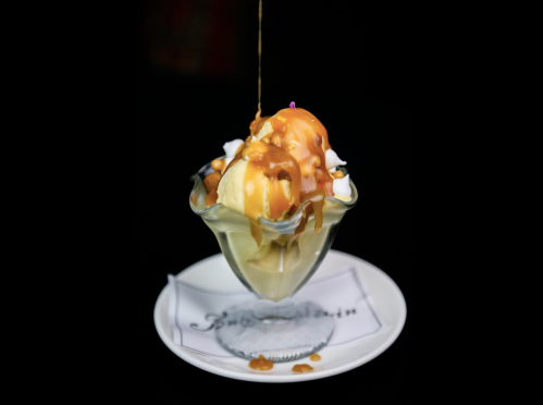 A glass dessert bowl filled with ice cream, topped with caramel sauce and nuts, on a white plate against a dark background.